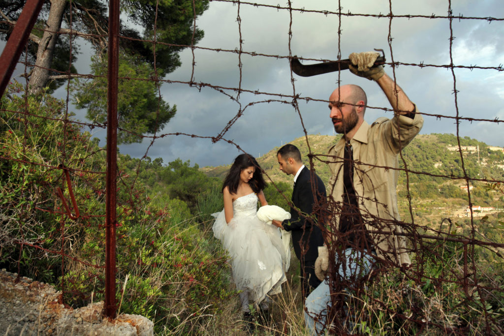 Figure 2. On the Bride’s Side / Io Sto con la Sposa, 2014, photo: Marco Garofalo. A rusty barbed-wire fence, embedded in crumbling concrete separates the viewer from hills covered in shrubs and a conifer in the foreground and the buildings of a distant city in the background. A pale-skinned person with a beard, shaved head, workers clothes and heavy gloves holds a machette and keeps watch over the hole they have cut in the fence as a bride and groom, young, olive-skinned, and slender and dressed in wedding formal wear help each other over the brush towards the gap. Storm clouds either gather or are being blown away.
