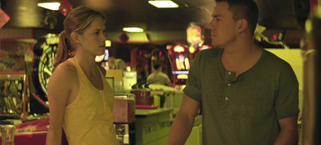 Figure 2. Brooke (Cody Horn) and Mike (Channing Tatum). Note the jaundiced color palette employed by Soderbergh in all scenes external to XQuisite. Magic Mike, author’s screenshot. Two white people face forward, with their heads turned towards each other. The scene is lit in sickly pale greens and yellows.