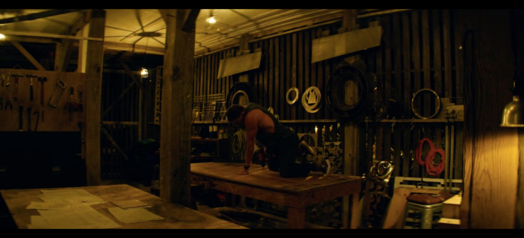 Figure 5. At this point, the drill appears to be animated by Mike’s thrusting pelvis. Magic Mike XXL, Warner Bros., 2015, author’s screenshot. In a workshop with large tables and a few tools a muscular man crouches on a tabletop, with a drill below his hips pointed towards the table's surface.