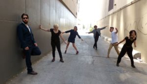 The six people from the first photo strike poses in an alley. Most of them have their arms spread; they are arranged at various distances towards the brightly-lit horizon at the end of the alley.