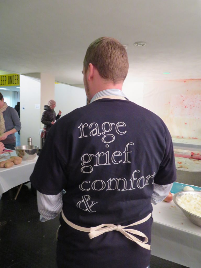 Back of a man wearing an apron and a shirt that says "rage grief comfort &", one word per line. There are people and tables in the background.