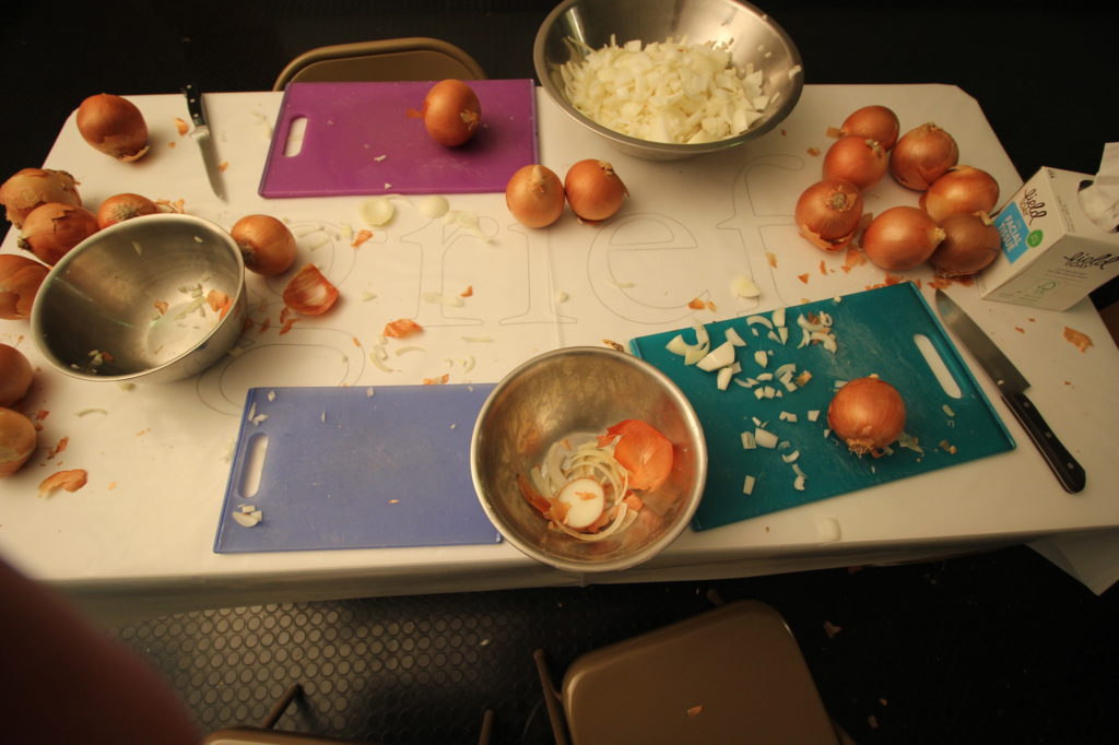Photo of a table with onions, bowls, knives, cutting boards, and tissues. Some of the onions are chopped.