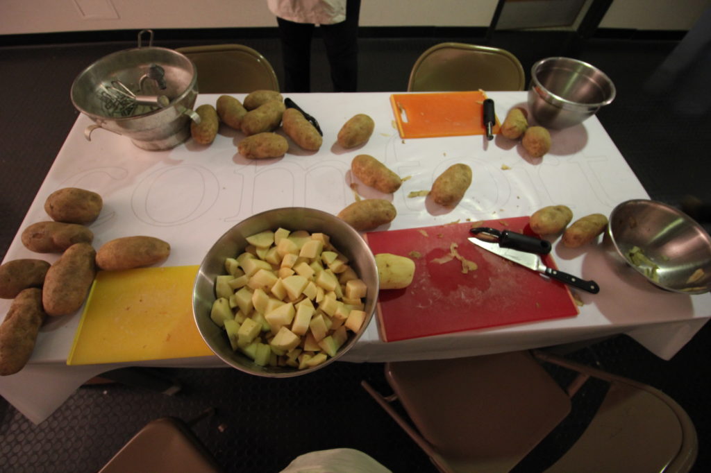 Photo of table with potatoes, bowls, knives, peelers, and cutting boards. Some of the tomatoes are cubed.