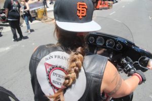 Fig. 3. Malone, Sheila. 2016 San Francisco PRIDE Parade Mindie’s Bike. We see the back of a motorcycle-riding, muscley woman wearing a "Dykes on Bikes" vest, braided hair, and a backwards baseball cap.