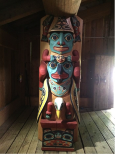 Figure 9: Interior of Main Dwelling of Eyak, Tlingit, Haida, Tsimshian Village Site, Alaska Native Heritage Center, Anchorage, AK. 2014. Photograph by Marina Tyquiengco. Inside the wooden room a tall, bright sculpture features many colorful faces arranged vertically, some human and some animal. Light spills through the open door.