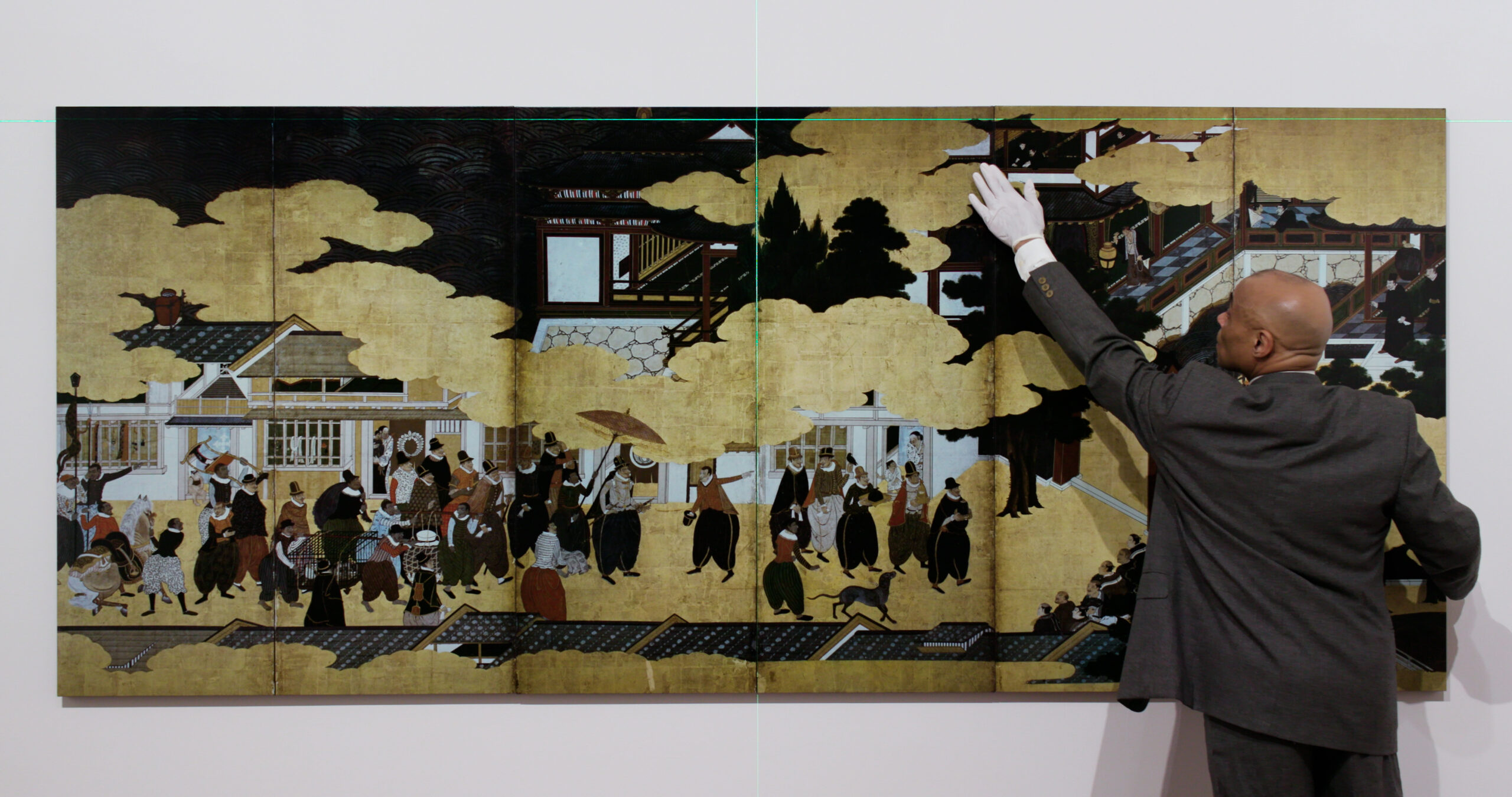 Peter Golightly in a suit, wearing a white glove on his left hand, is stretching his left arm to adjust the top right side of a painting, which consists of six panels. The painting is a photographic copy by Ikkō Narahara, who was asked to produce a collection of his photo works capturing a 15-century Japanese style work, which depicts a procession of non-Japanese foreign visitors.