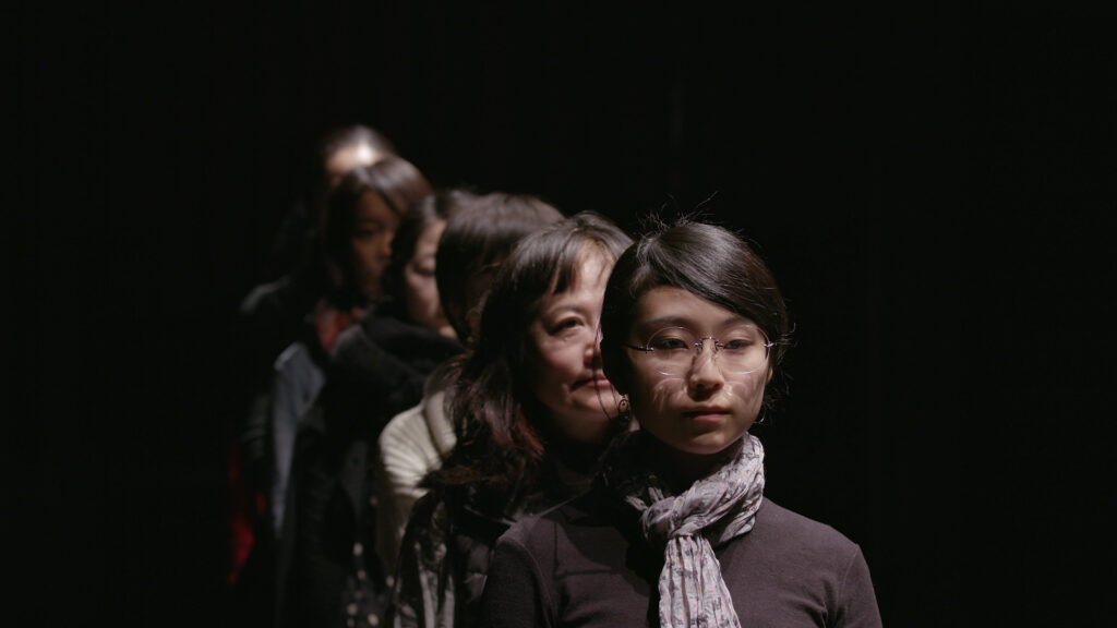 Six Asian women have lined up, facing forward, in a black, spartan, room.