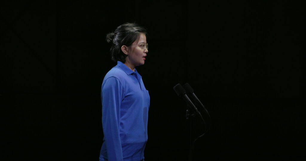 A Southeast Asian woman student wearing glasses and a blue shirt is standing in front of a mic, speaking.