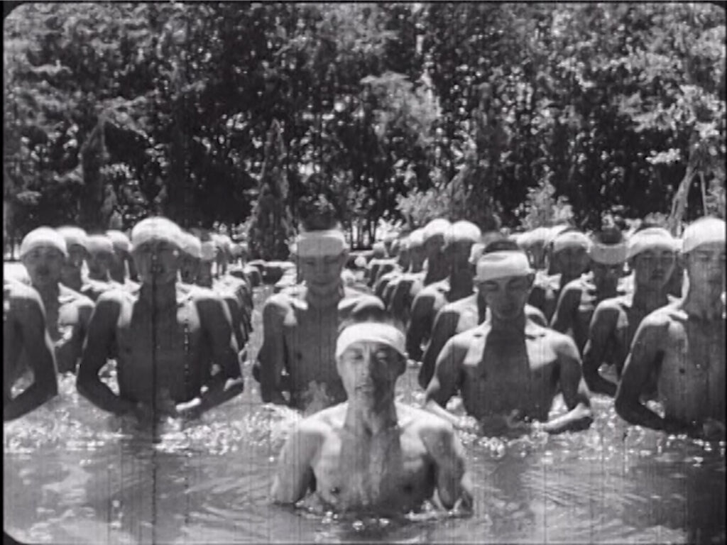 In a black-and-white picture, Taiwanese men, with their torsos bare while wearing white headbands, are standing in rows in a pool of water, forward facing toward the camera.