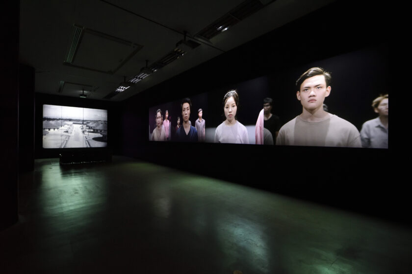 A pitch black gallery room, which shows film installations projected onto two walls. The installation on the right is a moving image of young refugees and immigrants in Japan, wearing simple pink, blue, or purple t-shirts and blouses, facing forward and looking at the camera. The image on the left is a black-and-white moving image showing rows of the training centers set up in Taiwan under Japanese colonial rule.