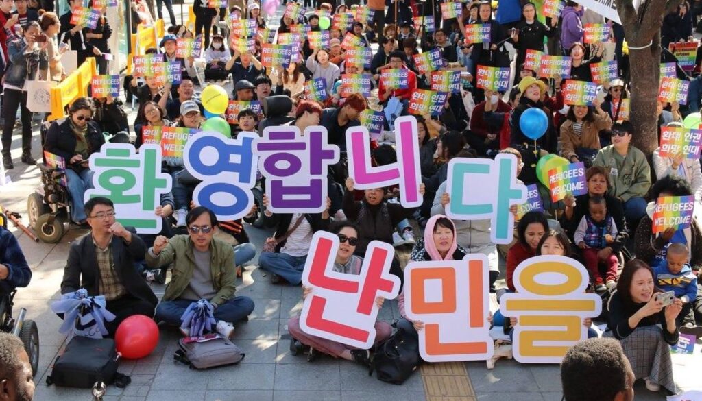 Picture of a multiethnic group of refugee rights activists in South Korea holding rainbow colored placards with a Korean and English message, “Refugees welcome.” At the front row, the rally participants are also holding large sign letters in rainbow colors, which say, “Refugees welcome.”