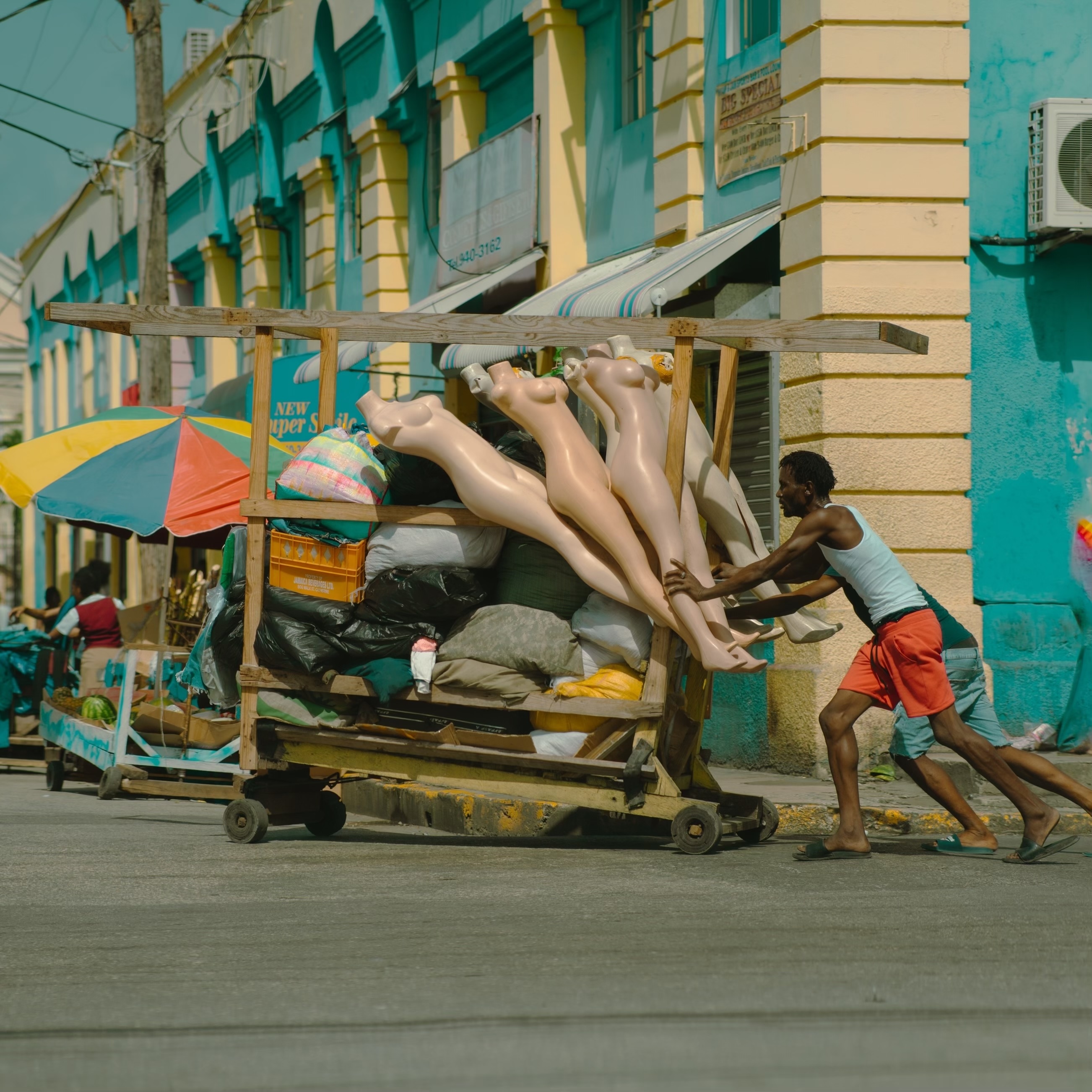 Two Black men push a handcart filled with white mannequins and other items down a busy street. A green and yellow building is in the background.