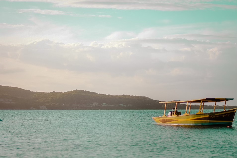 In the foreground a small boat painted black green and gold floats on a calm sea. In the distance is a mountain range. The entire image is covered in a pinkish haze.