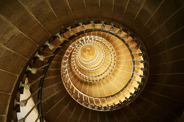 "un phare en coquille / Lighthouse like a shell" by TisseurDeToile. A photo of the spiral staircase of a lighthouse is shot from below. The stairs and railing spiral away into the light like a shell.