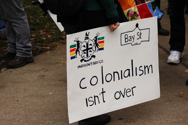 "Colonialism" by George Kourounis. Someone holds a handmade sign that reads "colonialism isn't over."