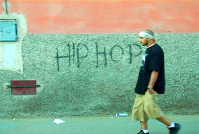 Still from "I Love Hip Hop in Morocco," by Joshua Asen and Jennifer Needleman, produced by Rizz Productions, Inc. 2007. A person, perhaps a Moroccan man, wearing sneakers, a bandana, baggy shorts and a long t-shirt, walks, hands in pockets, in front of a teal and pink wall graffitied with the words "HIP HOP."