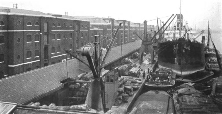 "The West India Dock, North Quay," from Owen Douglas's "London: The Port of the Empire" (1914), 34. A black and white photograph depicts a steam ship being loaded next to a row of brick factories on an urban dock side in the early 20th century.