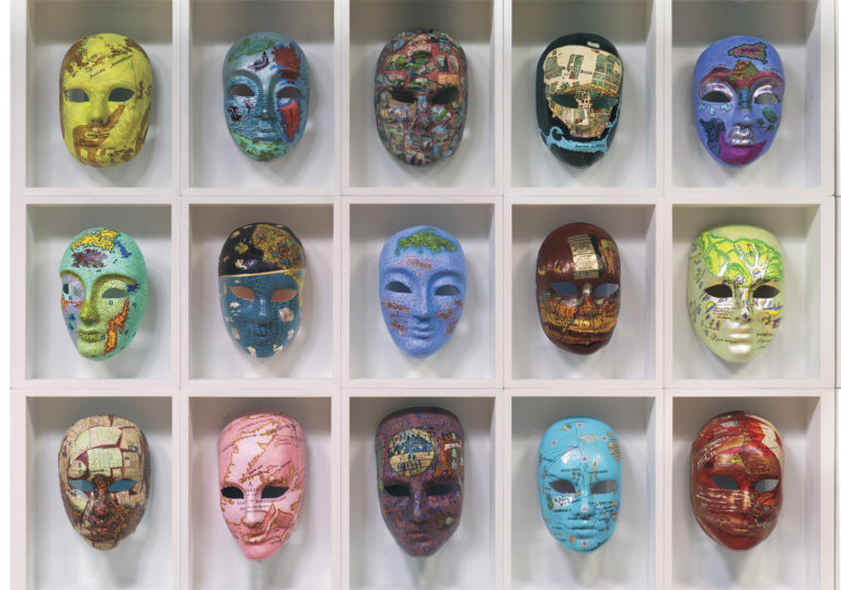 "Voyages, masks" by Joyce Kozloff. An image of colorful masks arranged in a grid of 5 by 3 on a white wall.