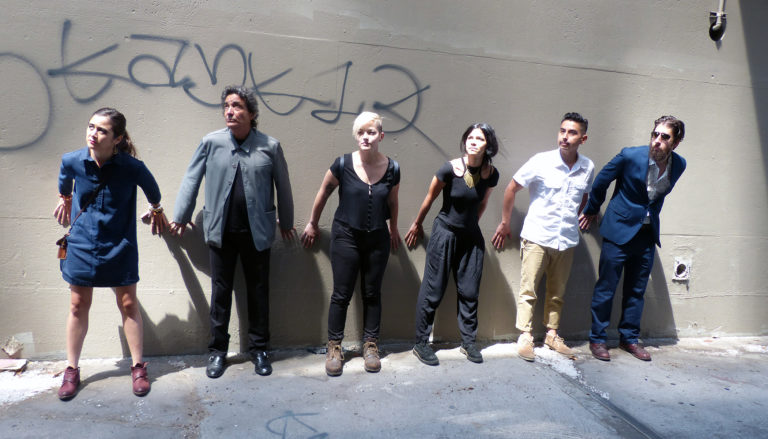 6 fashionably-dressed people stand in a line against a concrete wall. Their hands on the wall, they lean forward and look up and to the left.