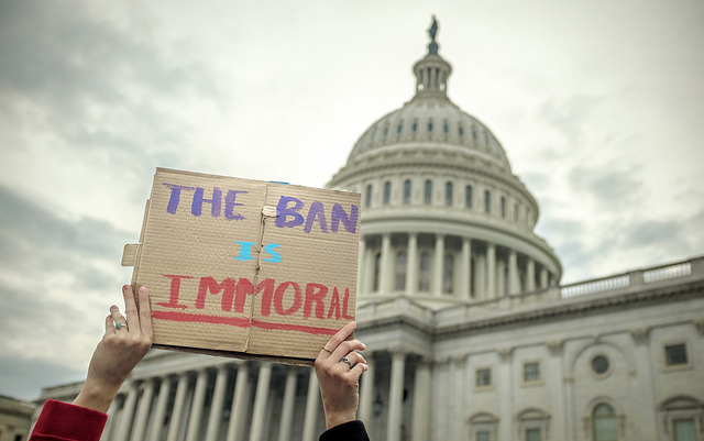 Photo of hands holding up a cardboard sign in from of the U.S. Capitol building. The sign says, "THE BAN IS IMMORAL."