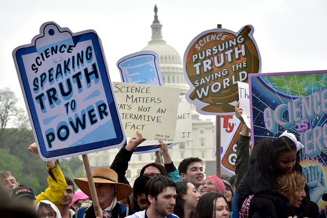 Photo of a protest with the U.S. Capitol building in the background. Various signs say "SCIENCE PURSUING TRUTH SAVING THE WORLD"