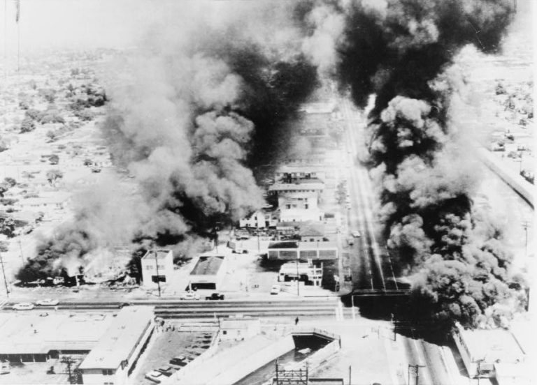 Black and white aerial photo of the Watts Rebellion in Los Angeles, showing smoke rising from several buildings.