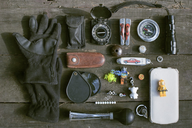 Survivalist items arranged on a wooden table. Items include gloves, compass, knife, flashlight, horn, and toys.