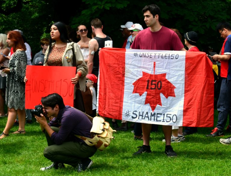 Protesters holding a Canada Flag upside down with #Unsettled150 and #Shame150 written on it