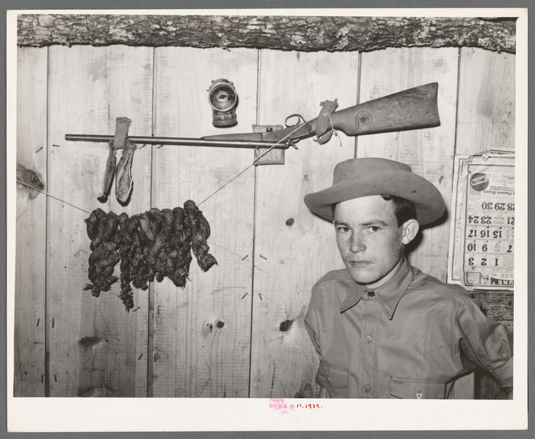 White teenager stands with his back against a wood-paneled wall. Hanging behind him are his twenty-two caliber rifle, a calendar, and home-cured tobacco.