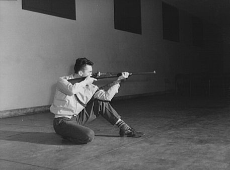 Black and white photo of a young white man with a rifle on his shoulder, crouching with his back to the exterior wall of a building and pointing the rifle into the darkness.