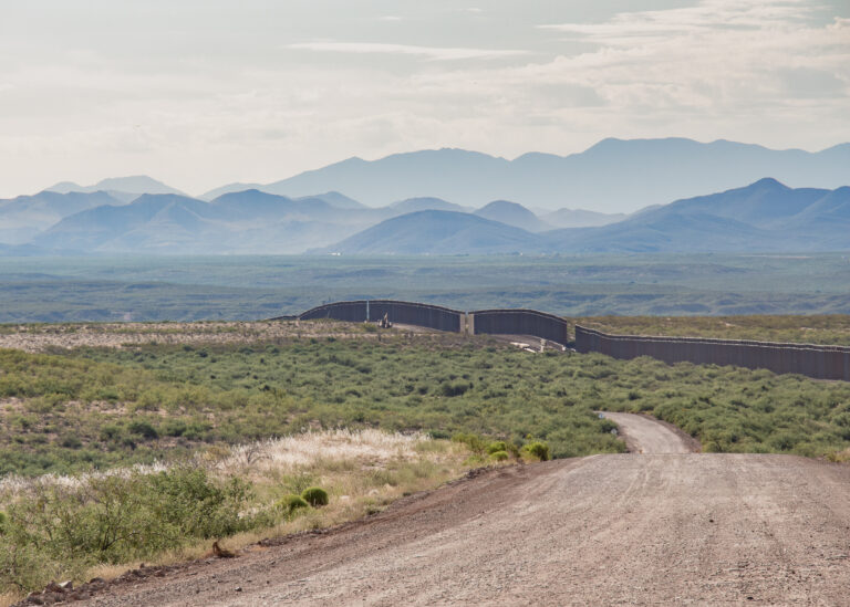 The image shows the border wall under construction in the mid-distance and rolling mountains on the other side.