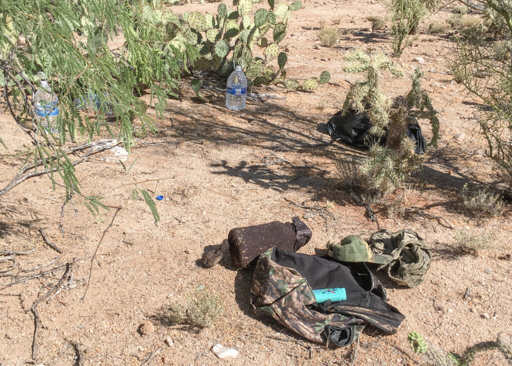 The image shows a used bottle of water, new bottles of water, and a backpack on the desert floor. 