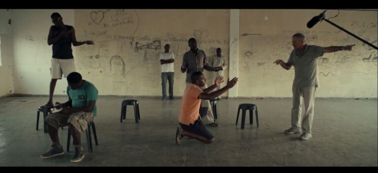 Still image of a performance. One man gestures with his arms out to another, who is pointing down, while one sits on a stool, another stands on a stool, and two stand against a graffitied wall in the backgroud.