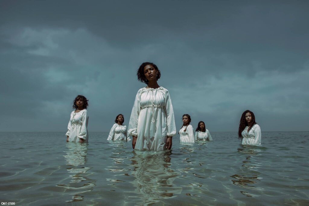 Six young women with dark skin dressed in white cotton dresses stand in a body of water, facing forwards in a group cluster.