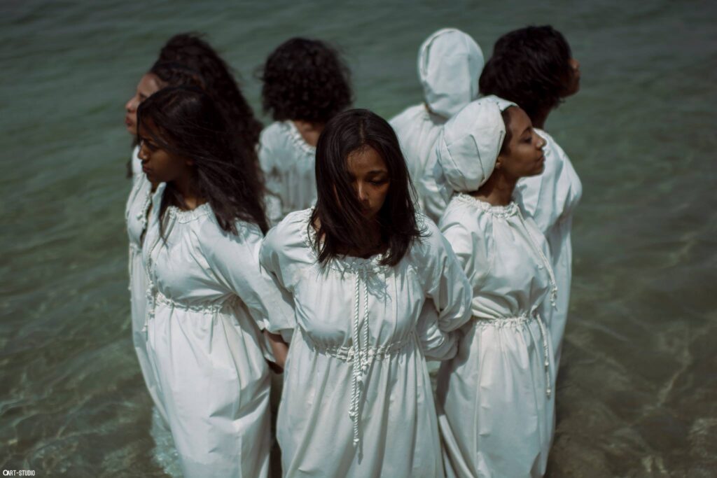 Seven young women with dark skin dressed in white cotton dresses stand in a body of water, locking elbows to form a tight circle while facing out.