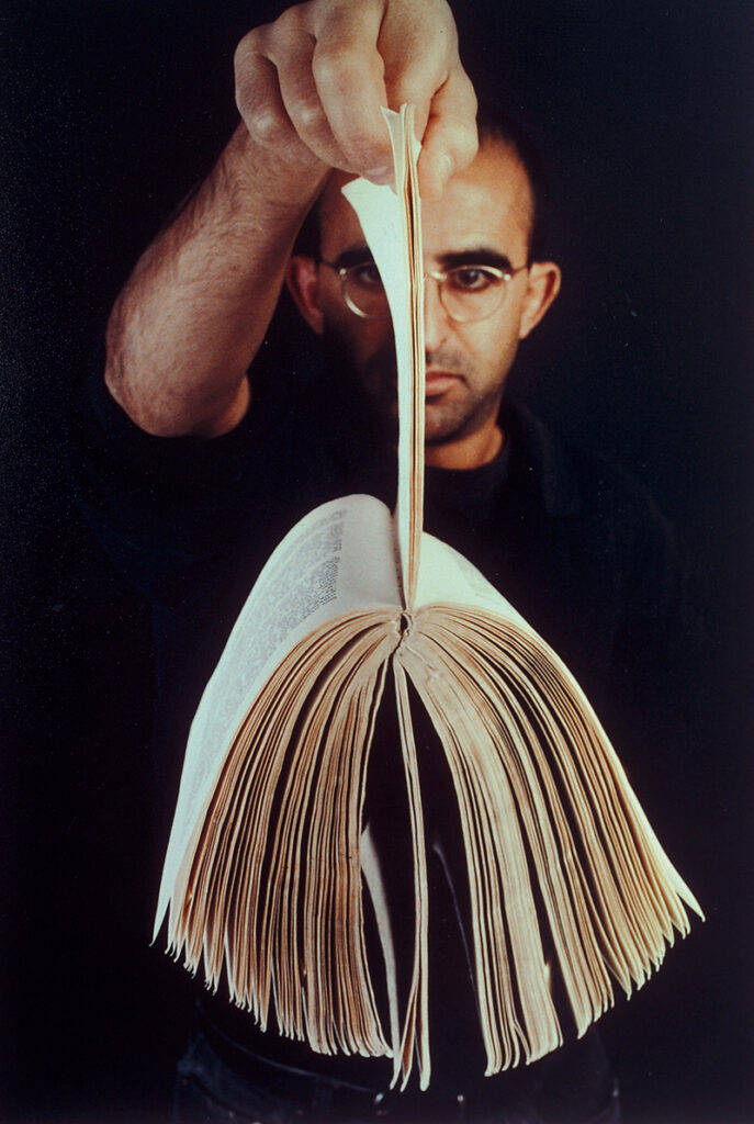 A man holding up a book with his right hand by a small number of pages so that the rest of the pages fan out to either side.