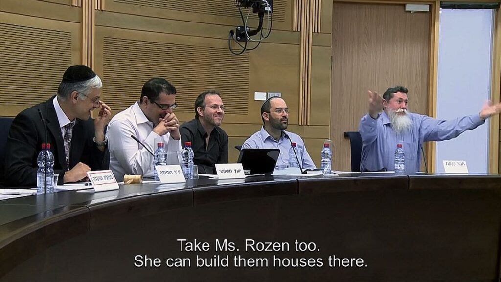 A group of men with microphones and name plates sitting behind a table. One gestures with his hands while the others laugh. A caption reads, "Take Ms. Rozen too. She can build them houses there."