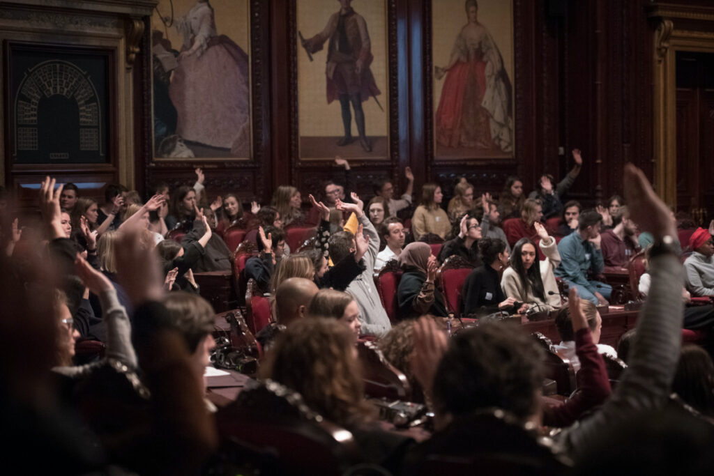Photo of a performance of De Waarheidscommissie by Kurt Van der Elst. Members of the audience raise their hands.