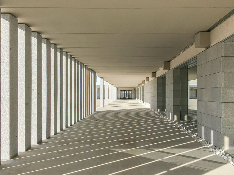 Color photo of La Academia de Oficiales de la Guardia Civil in Aranjuez, Spain. Long passageway with rectangular pillars on the side. The light passing through the pillars makes a checkered pattern on the floor.