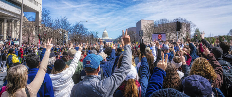 A close up image of a large crowd outside holding up the peace sign. In the center at a far distance, is the US Capital Building and to the right is a large mega screen projecting the image of the speaker: X González.
