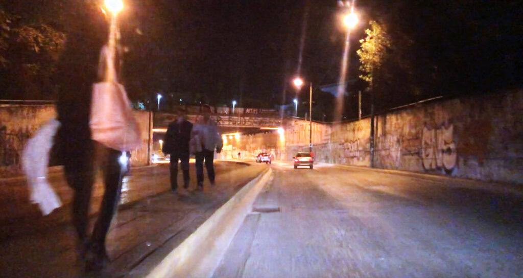 Photograph from a moving car, which depicts the underground passage of Moustoxidi street with people walking on the median strip during night.