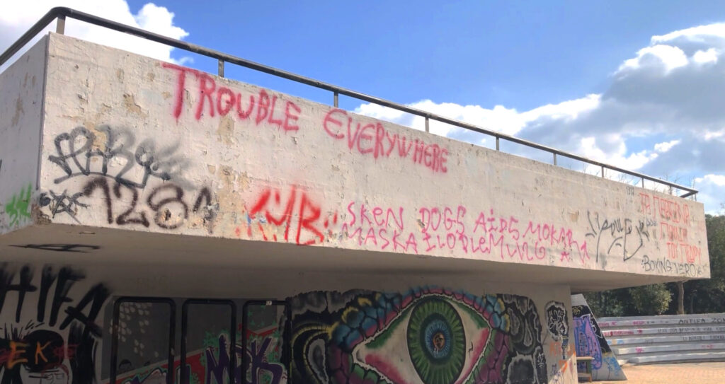 Photograph of a cement wall full of graffiti pieces and tags, next to an amphitheatre. At the bottom of the wall stands out a piece representing an eye, while on the top of the wall, among several tags, there is the phrase “Trouble Everywhere” written in red paint.