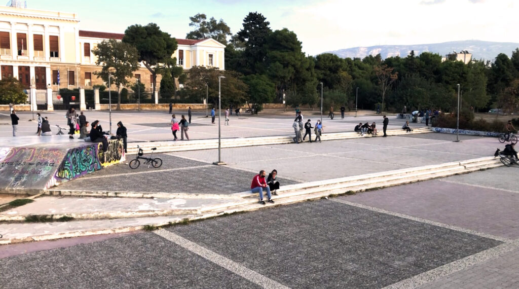 Photograph of the open space in the middle of the square and in front of the neoclassical building of the Hellenic National Defence College. In the photograph we see small groups of people and couples hanging out in the afternoon.