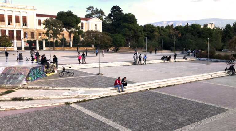 Photograph of the open space in the middle of Protomagias Square and in front of the neoclassical building of the Hellenic National Defence College. In the photograph, small groups of people and couples hanging out in the afternoon.