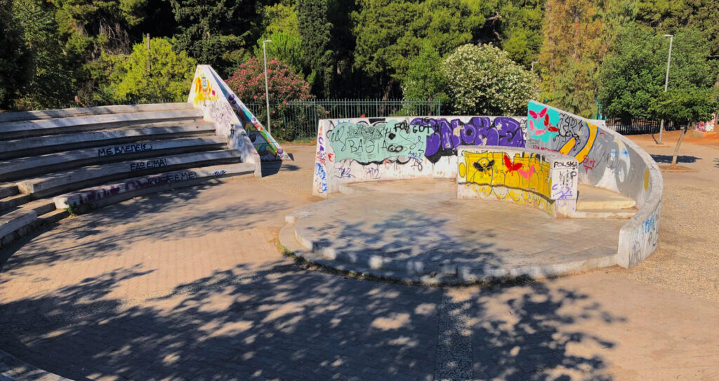 Photograph of the amphitheatre early in the morning. It depicts graffiti painted walls, while shadows from the surrounding trees fall on the amphitheatre at the centre of the picture.