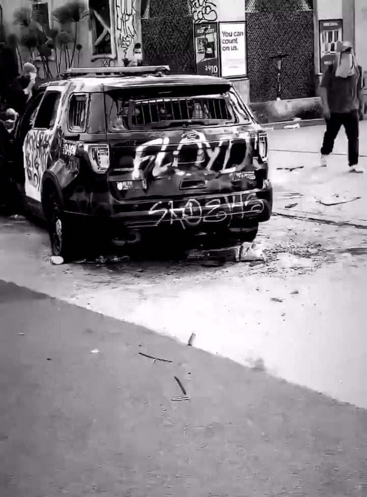 A burnt and smashed police car that is tagged with the graffiti 'FLOYD'