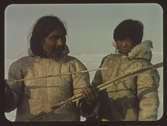 Color photograph of a Netsilik man and boy. The man is showing the boy some arrows that he is holding in his hands.