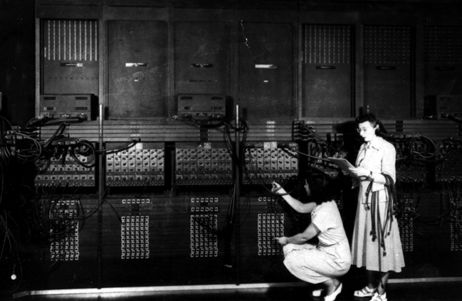 Two women in white dresses hold wires and a notebook, while working on the enormous ENIAC computer- which takes up the entire wall in behind them.