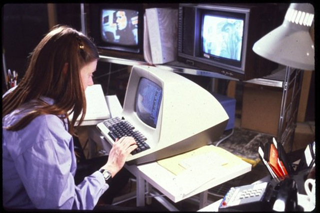 A woman types on a computer keyboard, with two TV screens in the background. A telephone, desk lamp, and office supplies sit on her desk.