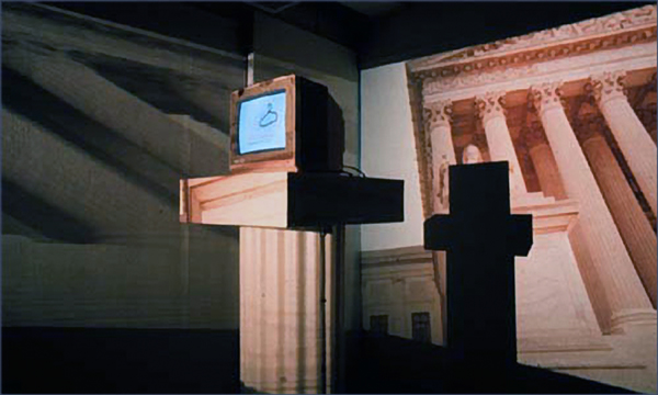 A computer with a wooded frame sits atop a pedestal. A pillared building is projected in the right-hand background; the same image is projected to the left, but is distorted.